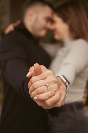 Close-up shot of clasped hands showing an engagement ring with two people embracing in soft focus background.