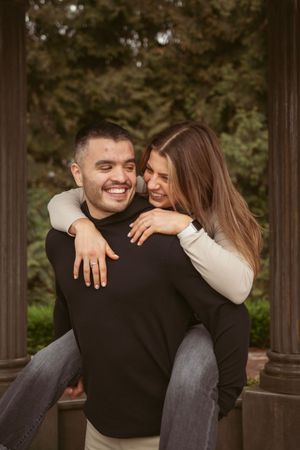 A happy couple embraces outdoors, with her giving him a playful piggyback ride against a natural background.