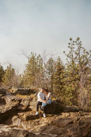 Two people sitting close together on rocky terrain with pine trees in the background.