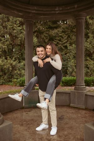 Playful couple sharing a piggyback moment in a garden gazebo surrounded by lush greenery.