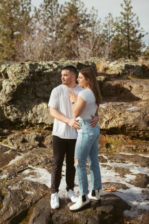 A romantic couple walks together on rocky terrain near evergreen trees.
