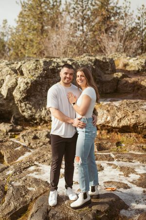 Engaged couple embracing on rocky terrain with winter forest backdrop.