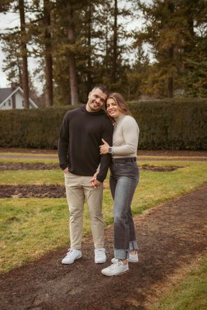 Happy couple in casual attire posing together on a path with evergreen trees in background.