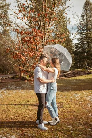Couple sharing romantic moment under clear umbrella in autumn forest setting with orange foliage.