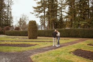 A person stands on a path in a garden with tall evergreen trees and manicured hedges in the background.