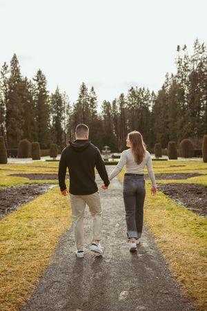 Two people holding hands while walking down a gravel path flanked by evergreen trees.
