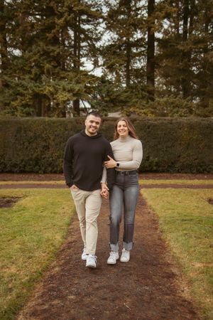 A couple walks together holding hands on a dirt path surrounded by evergreen trees and manicured hedges.