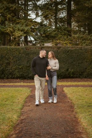 Two people walk hand in hand together along a scenic autumn garden pathway lined with lush green hedges.