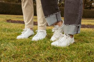 Two pairs of white sneakers on grass with rolled up jeans and khaki pants.