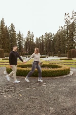 Two people playfully running together on a curved gravel path surrounded by evergreen trees and manicured hedges.