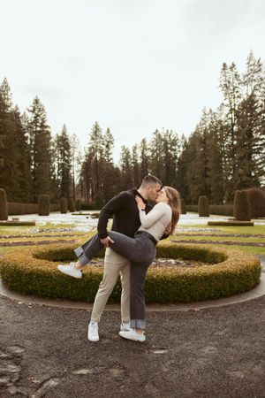 A couple embraces in a romantic dip pose in front of ornate garden hedges and tall evergreen trees at dusk.