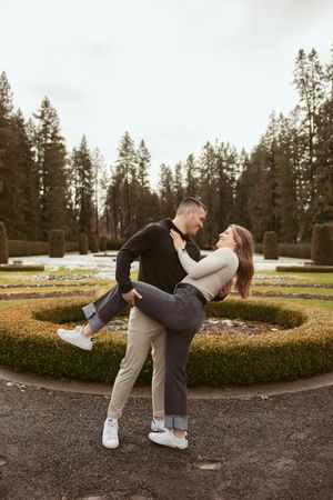 Playful couple dancing together in a formal garden with evergreen trees in background.