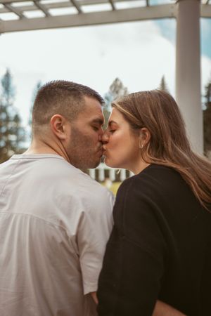 A couple in white and black attire sharing a romantic kiss under a covered porch.