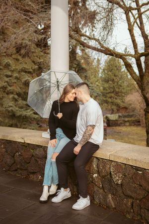 Couple sharing romantic moment on stone wall bench with clear umbrella and trees in background.