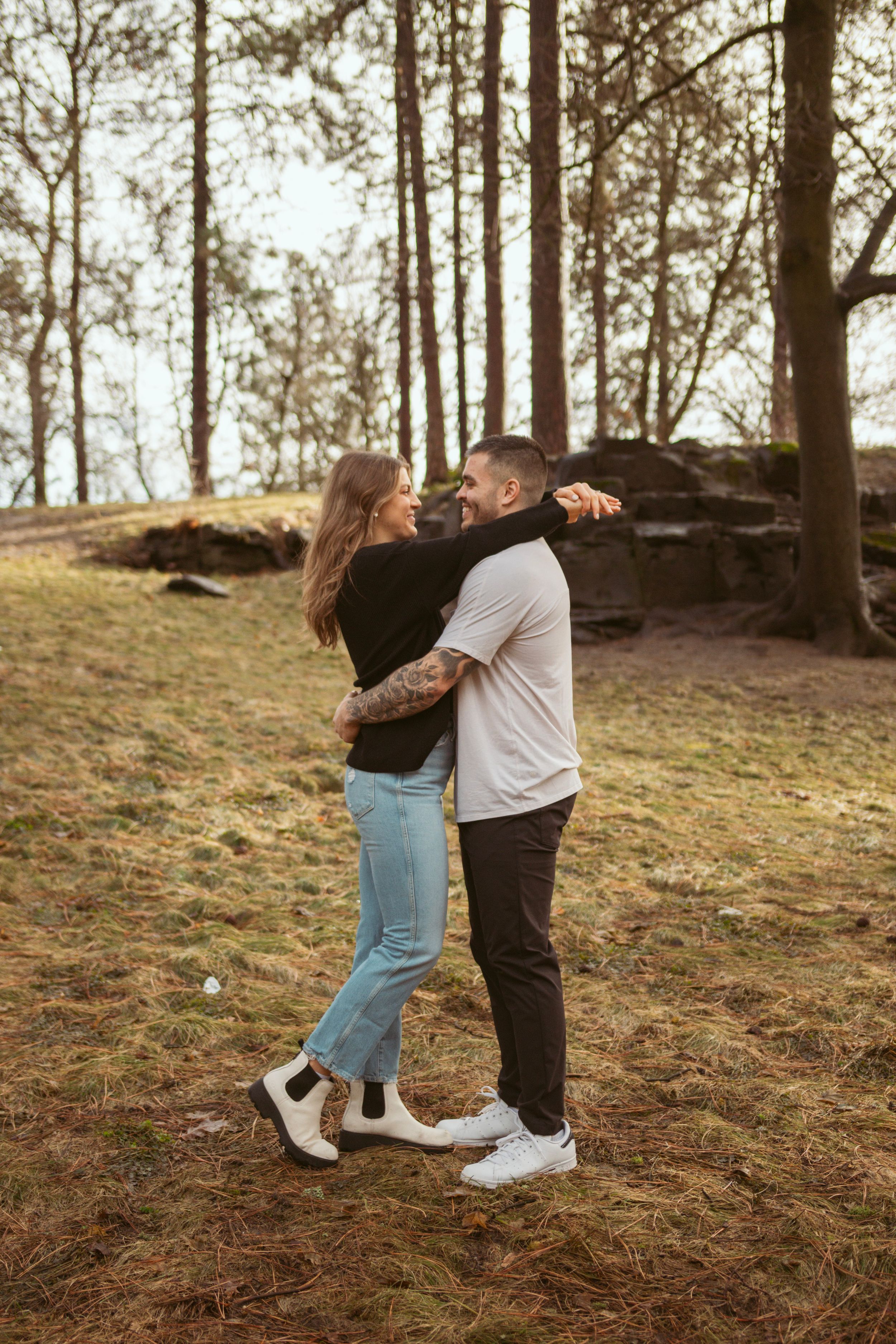 Two people share a romantic dance together on a hillside surrounded by tall pine trees at sunset.