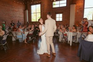 Guests watch a couple dance in a rustic reception venue with exposed brick walls and string lights.