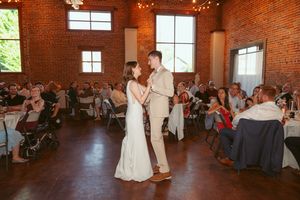 Couples dancing at a wedding reception in an industrial brick venue with large windows and decorative lighting.
