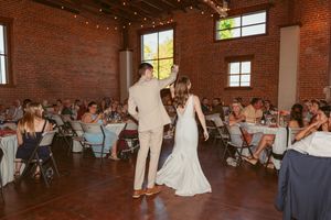 Two people dancing at a wedding reception in a rustic brick venue with string lights and guests watching.