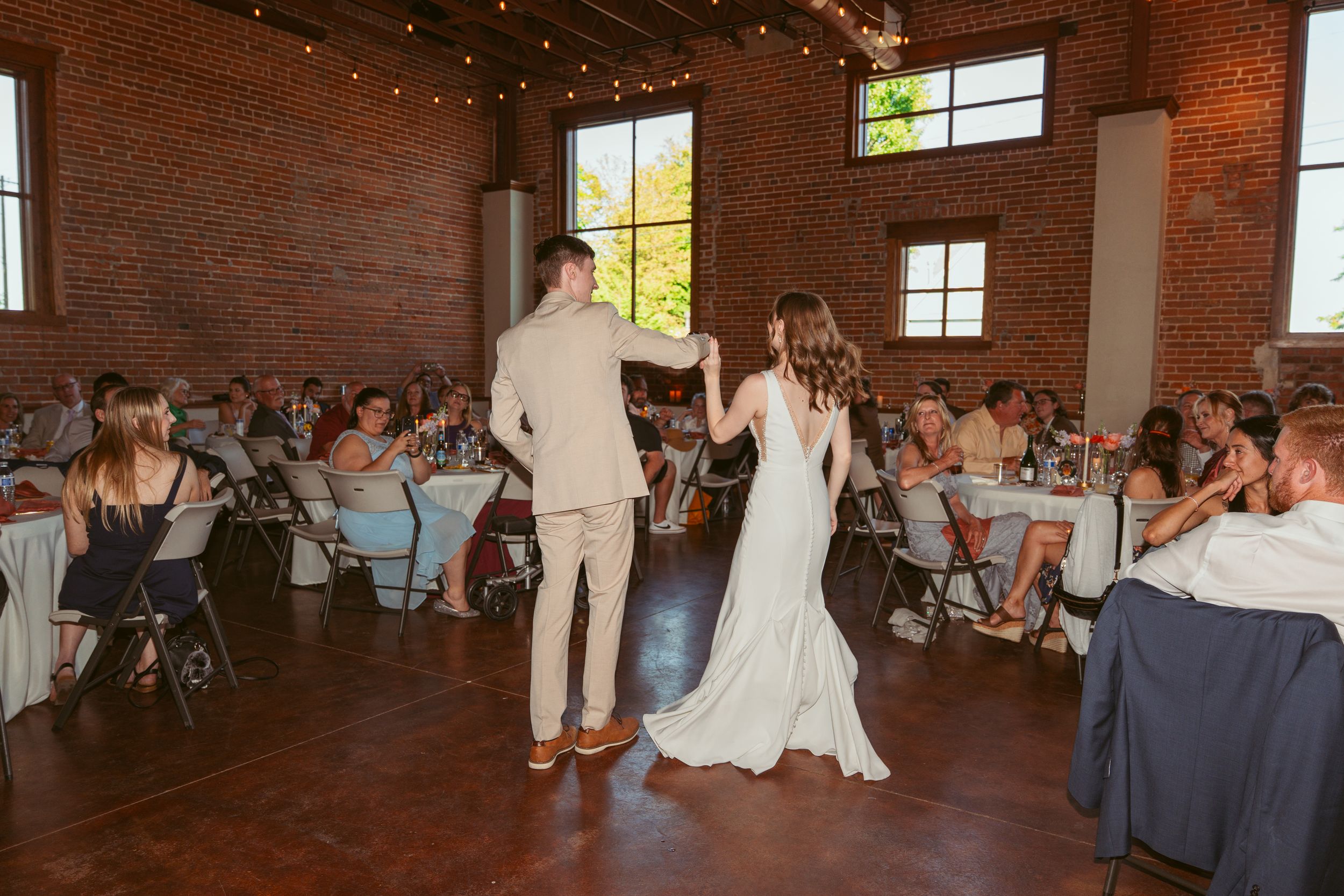 A couple performs their first dance at a rustic wedding reception venue with exposed brick walls and golden sunlight.