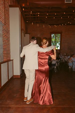 Two dancers in elegant attire share an intimate moment during a reception in a rustic brick-walled venue with string lights.