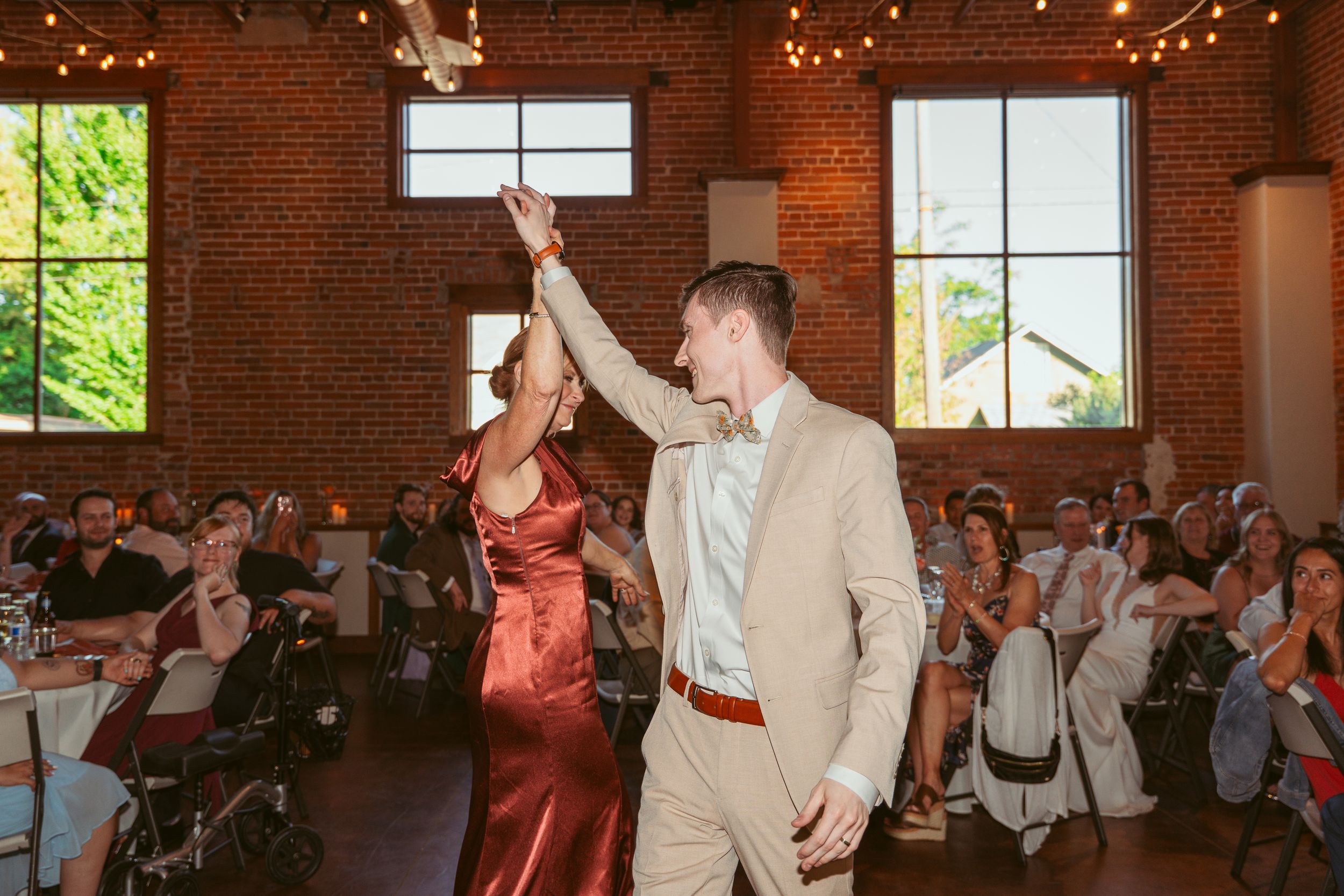 Two guests celebrating with raised hands during a wedding reception in a brick-walled venue with string lights.