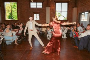 Dancers in formal attire perform an energetic dance routine on a hardwood floor at a reception venue.