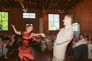 Couple in red dress and beige suit dancing energetically at a wedding reception in an exposed brick venue.