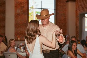 A couple dances at a wedding reception in a rustic brick venue with guests seated at tables in the background.