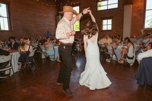 Wedding couple dancing while guests seated at round tables watch in a brick-walled reception space.