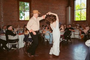 Two celebrants share a lively swing dance in a rustic brick venue with guests watching from folding chairs.