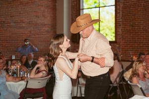 A dance takes place at a wedding reception in a rustic brick venue with natural light streaming through windows.