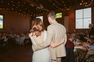 A couple stands together at their reception while guests seated at tables look on in a brick-walled venue.
