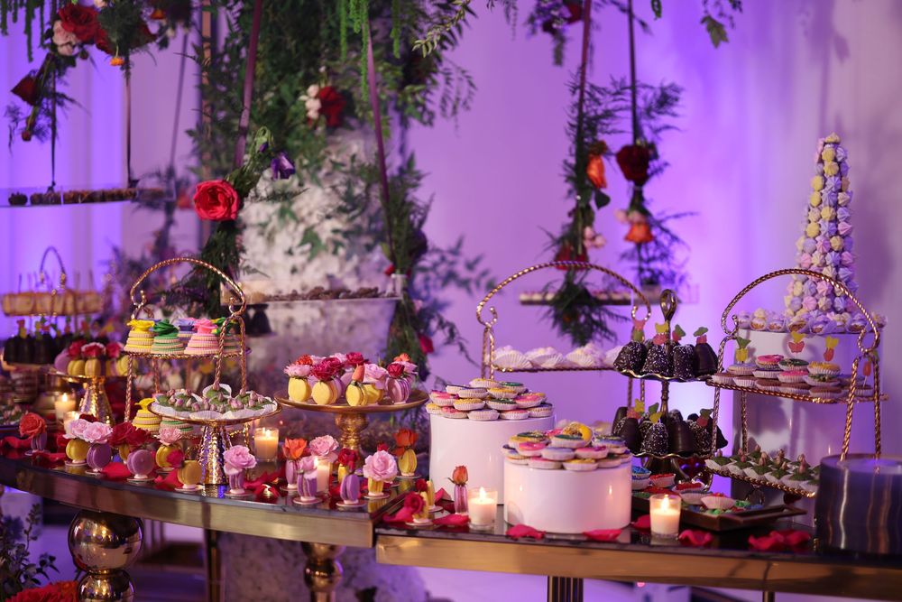 A luxurious dessert table decorated with elegant tiered displays, flowers, and treats illuminated by purple lighting.