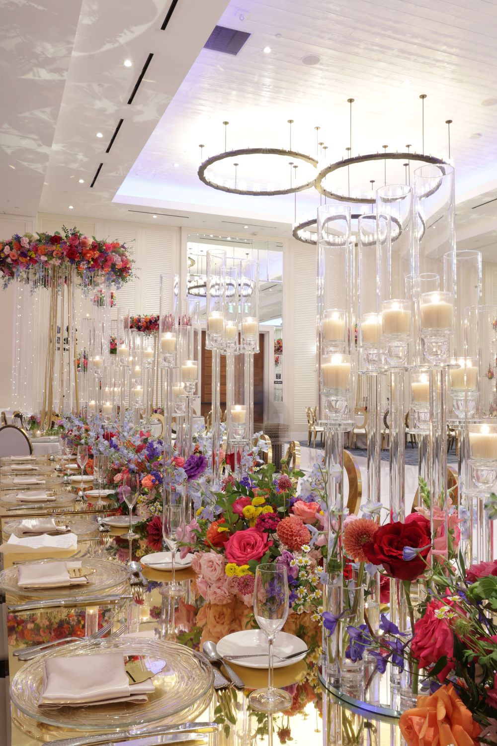 Elegant wedding reception table with crystal chandeliers and vibrant floral centerpieces featuring red and pink blooms.