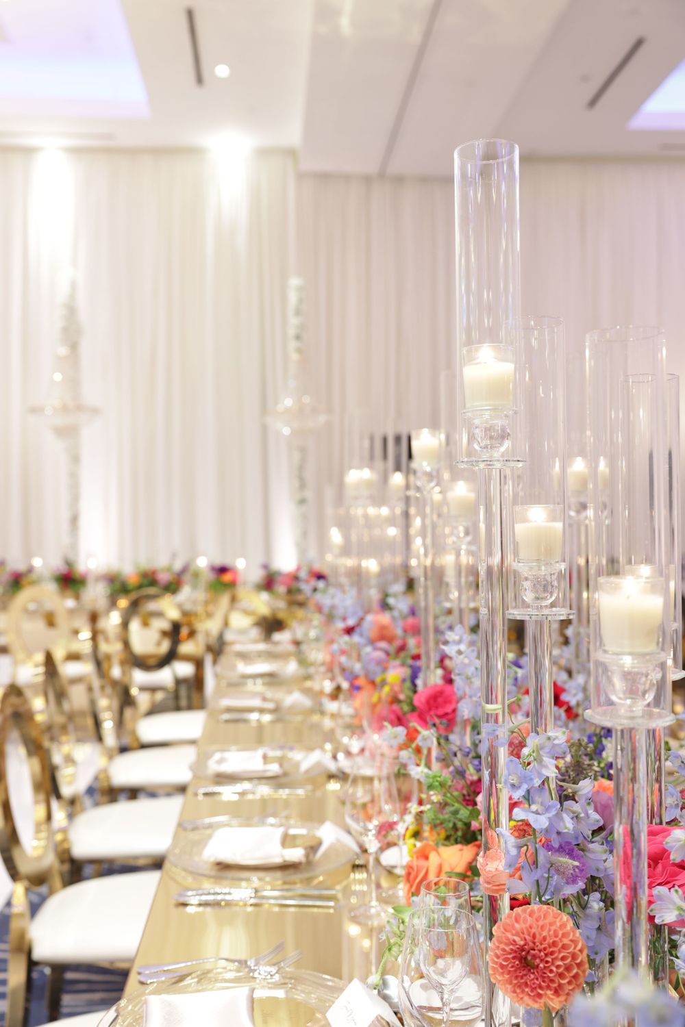 Elegant wedding reception table with tall glass candle holders, colorful floral arrangements and gold chiavari chairs.