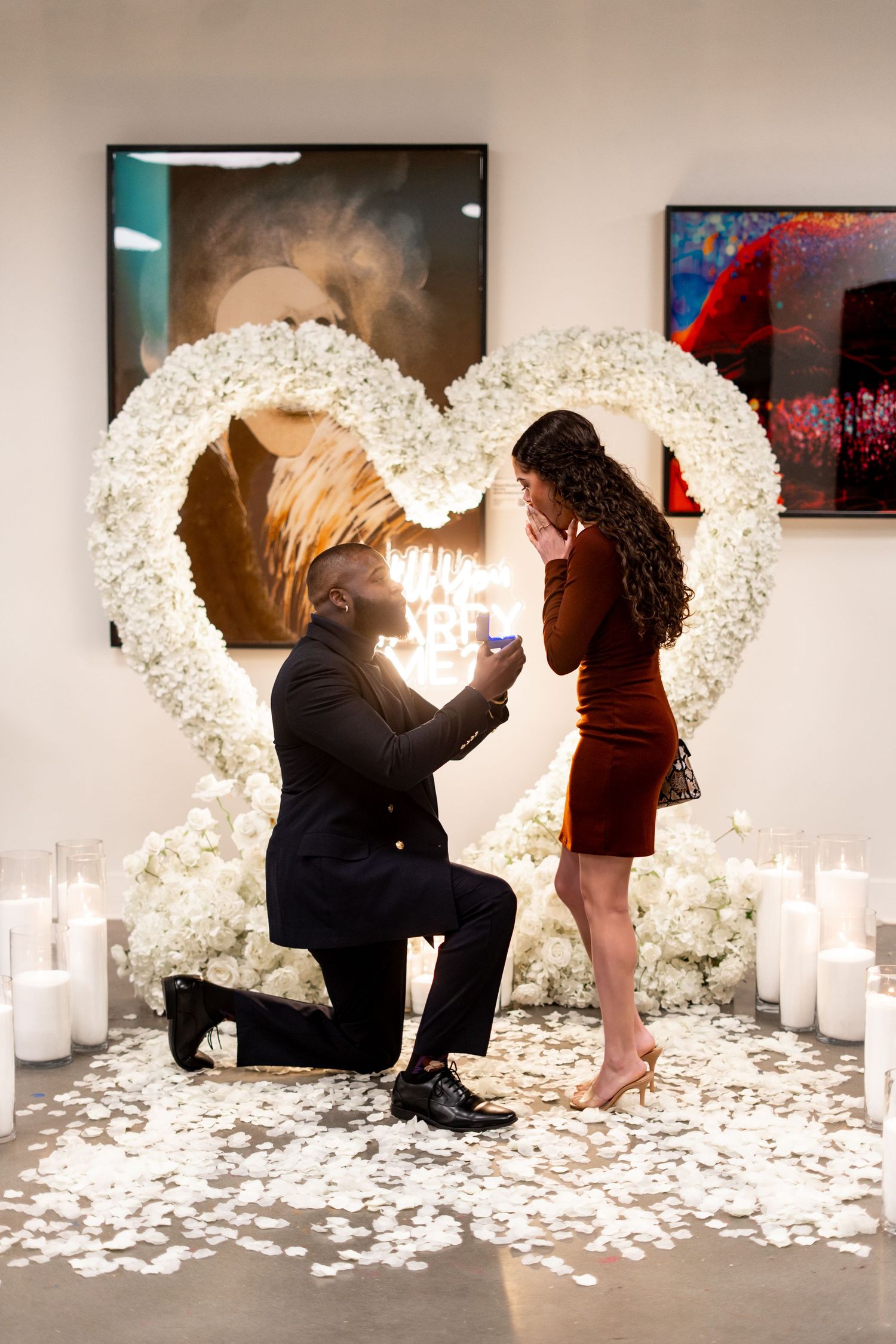 A romantic proposal scene with a large illuminated white heart arch and candles creating an intimate atmosphere in an art gallery.