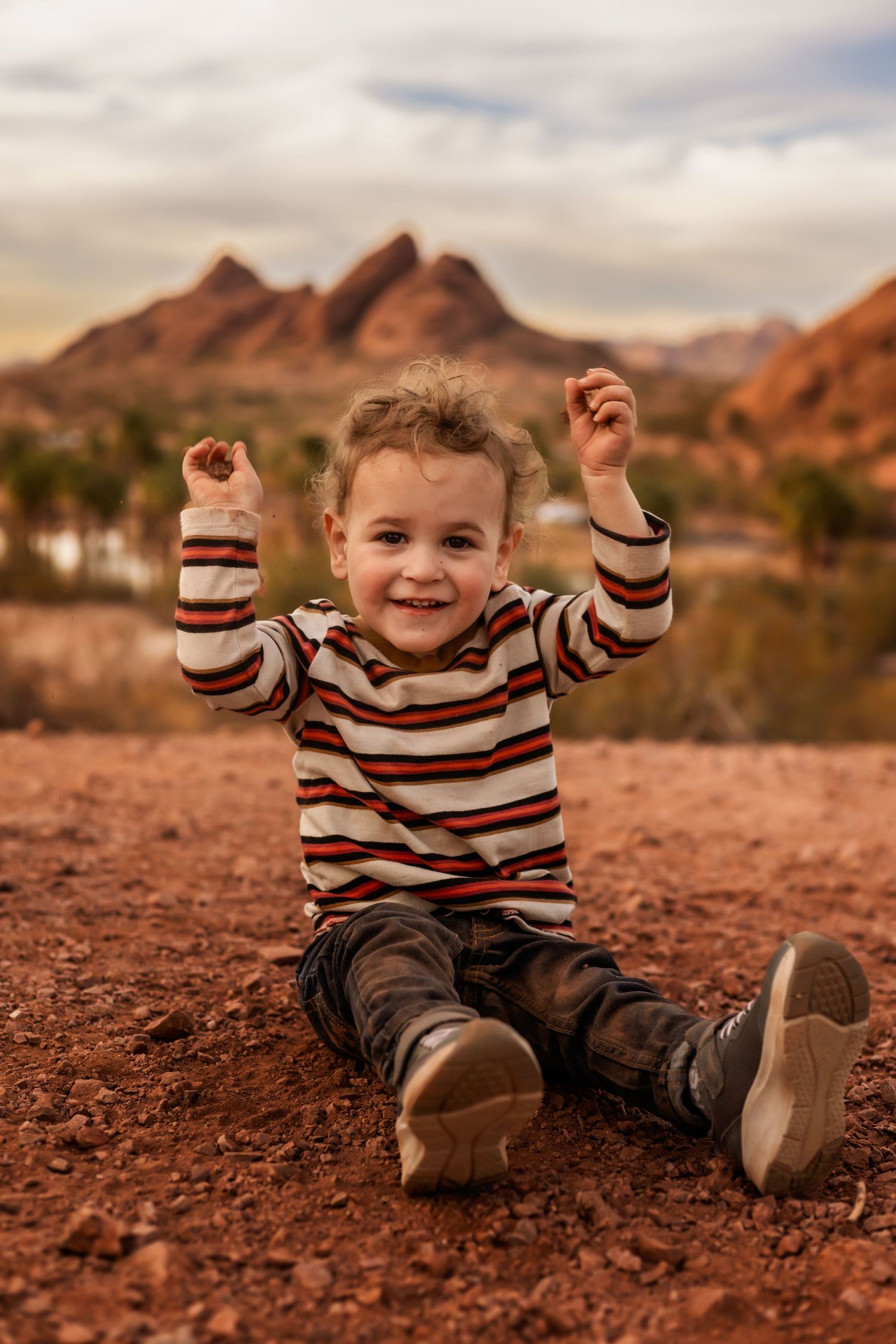 Joyful small child in striped sweater raises arms while sitting on red desert ground with mountain peaks in background.