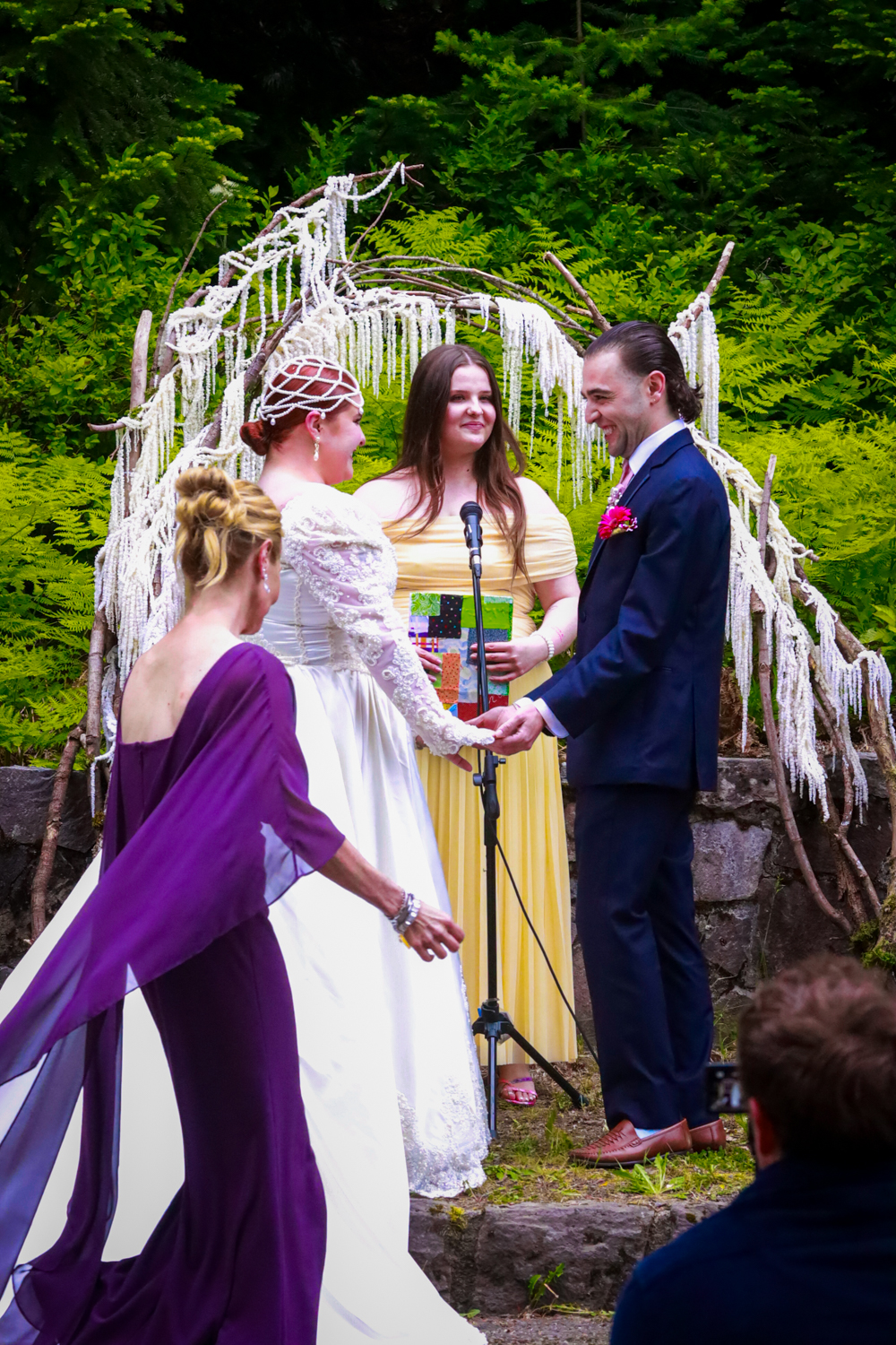 Outdoor wedding ceremony under a rustic white arch decorated with fabric and flowers in a lush garden setting.
