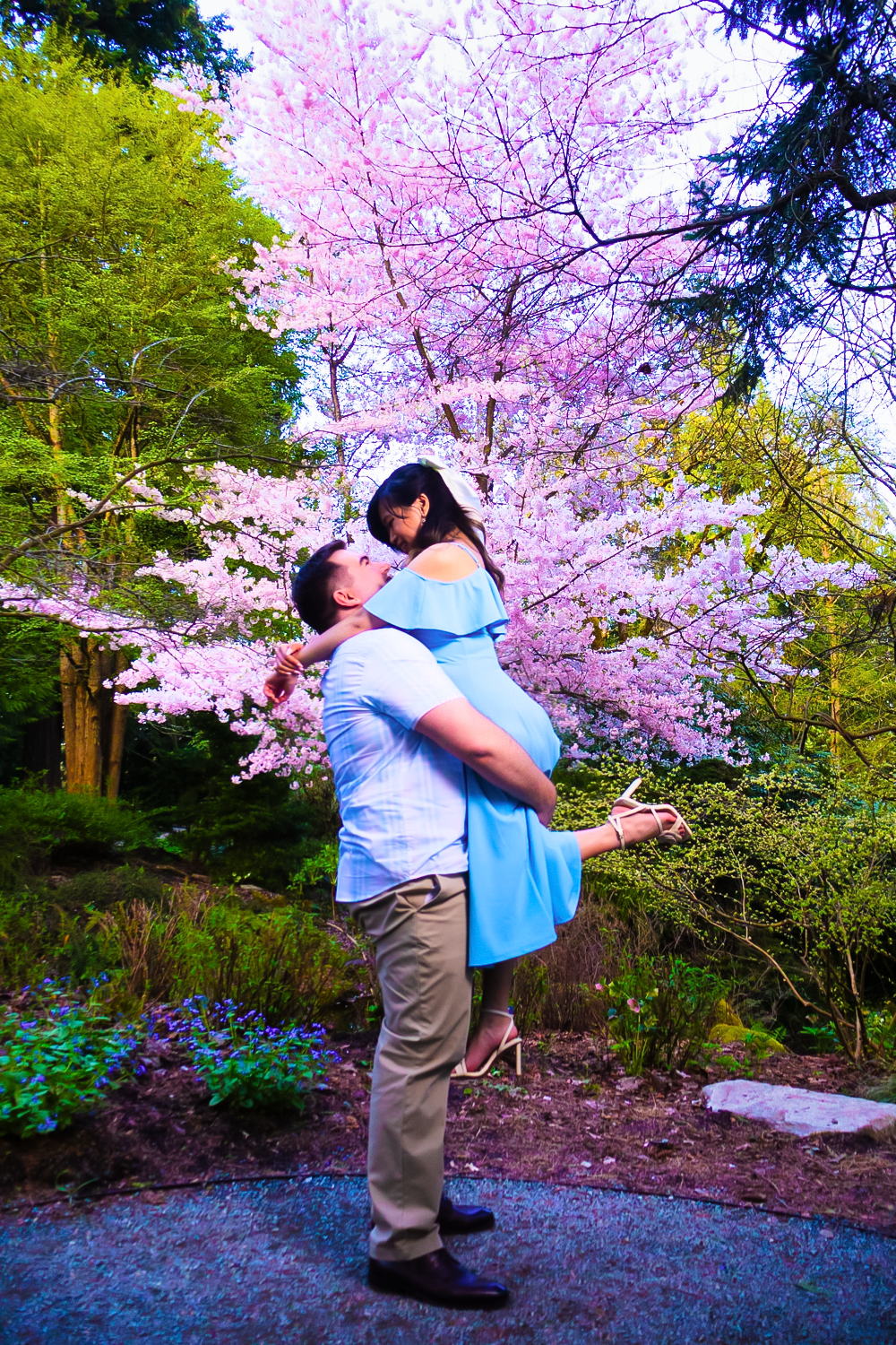 A couple embraces on a garden path surrounded by vibrant azalea flowers and lush greenery.