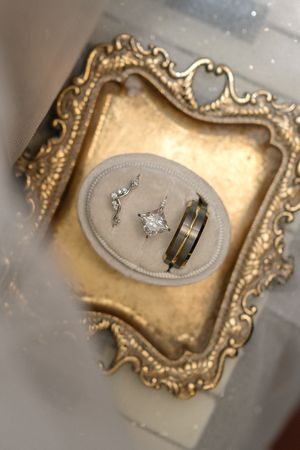 Wedding rings and jewelry displayed in an ornate vintage gold frame on a velvet cushion with soft focus background.