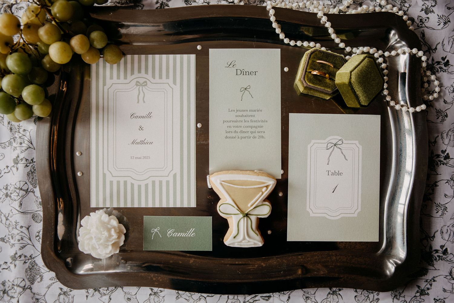 Elegant wedding stationery flat lay on silver tray with grapes, pearls, and green velvet ring box.