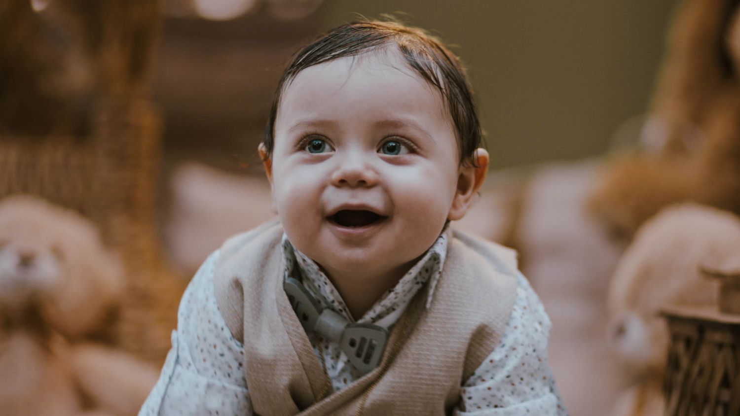 A baby in a light colored sweater smiles brightly while crawling on the floor.