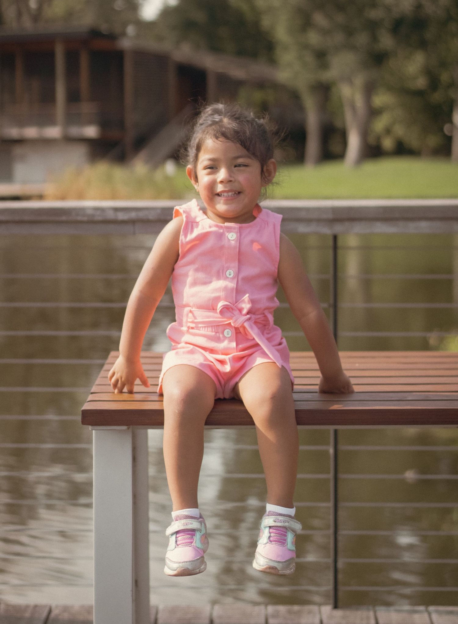 Smiling toddler girl in pink romper sitting on wooden bench by a lake on a sunny day.