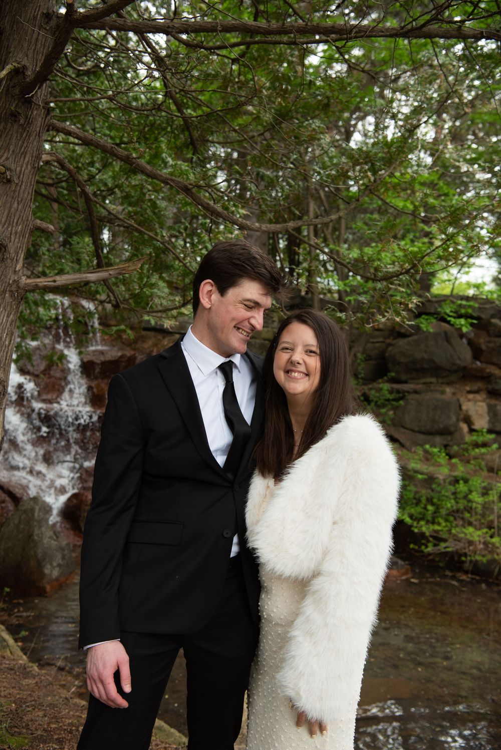 A couple shares a joyful moment together in formal attire by a waterfall and stone wall in a forest setting.