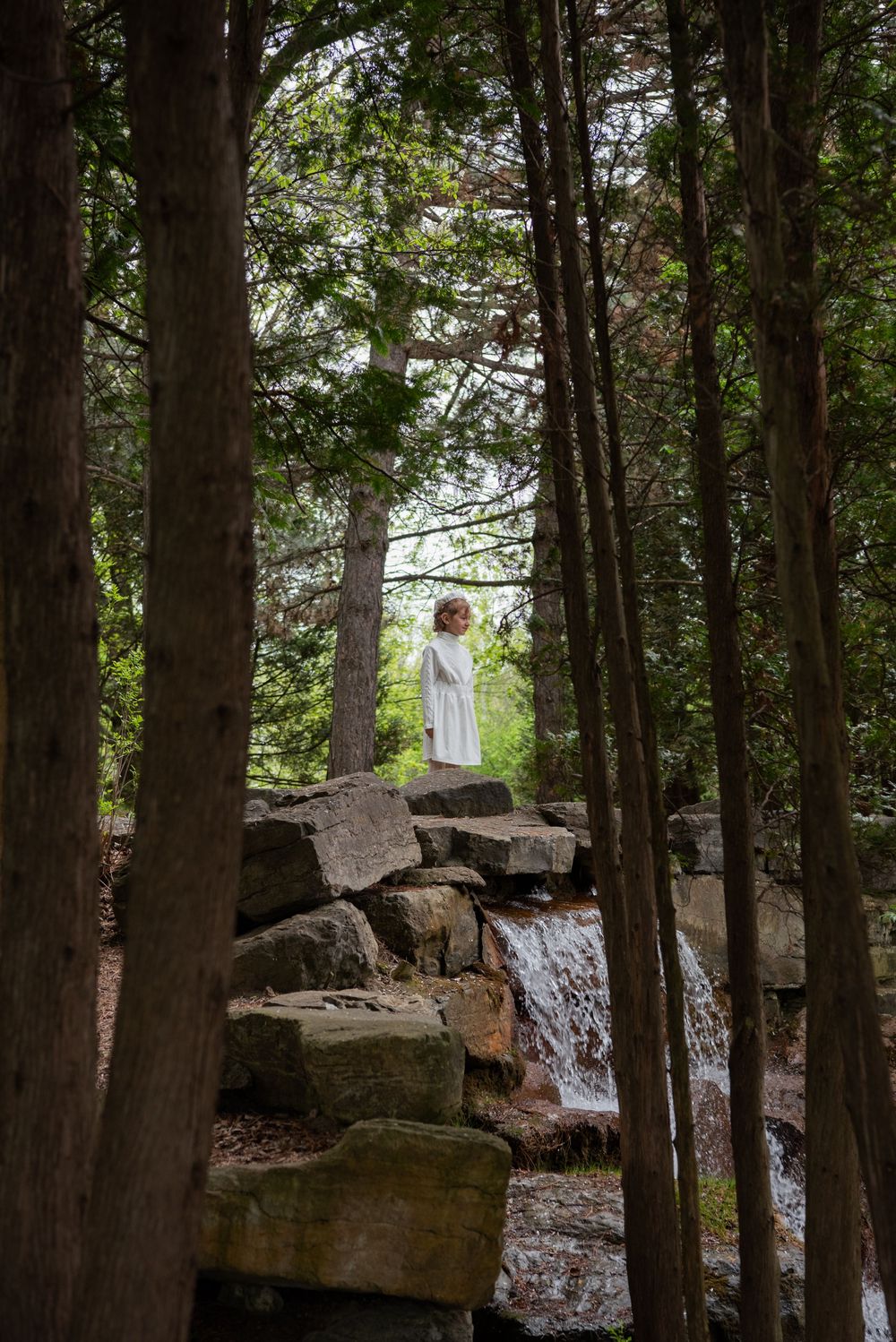White-clad figure stands atop rocky waterfall formation in a serene wooded setting.
