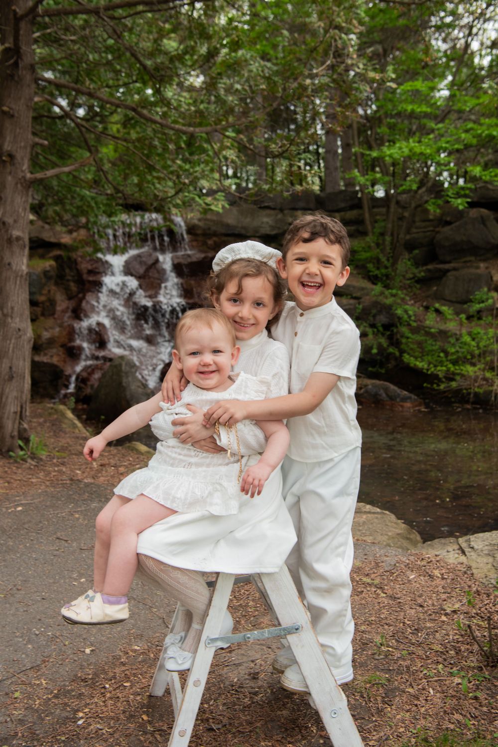 Three children in white outfits sit and stand together on a stool with a waterfall cascading in the forest background.