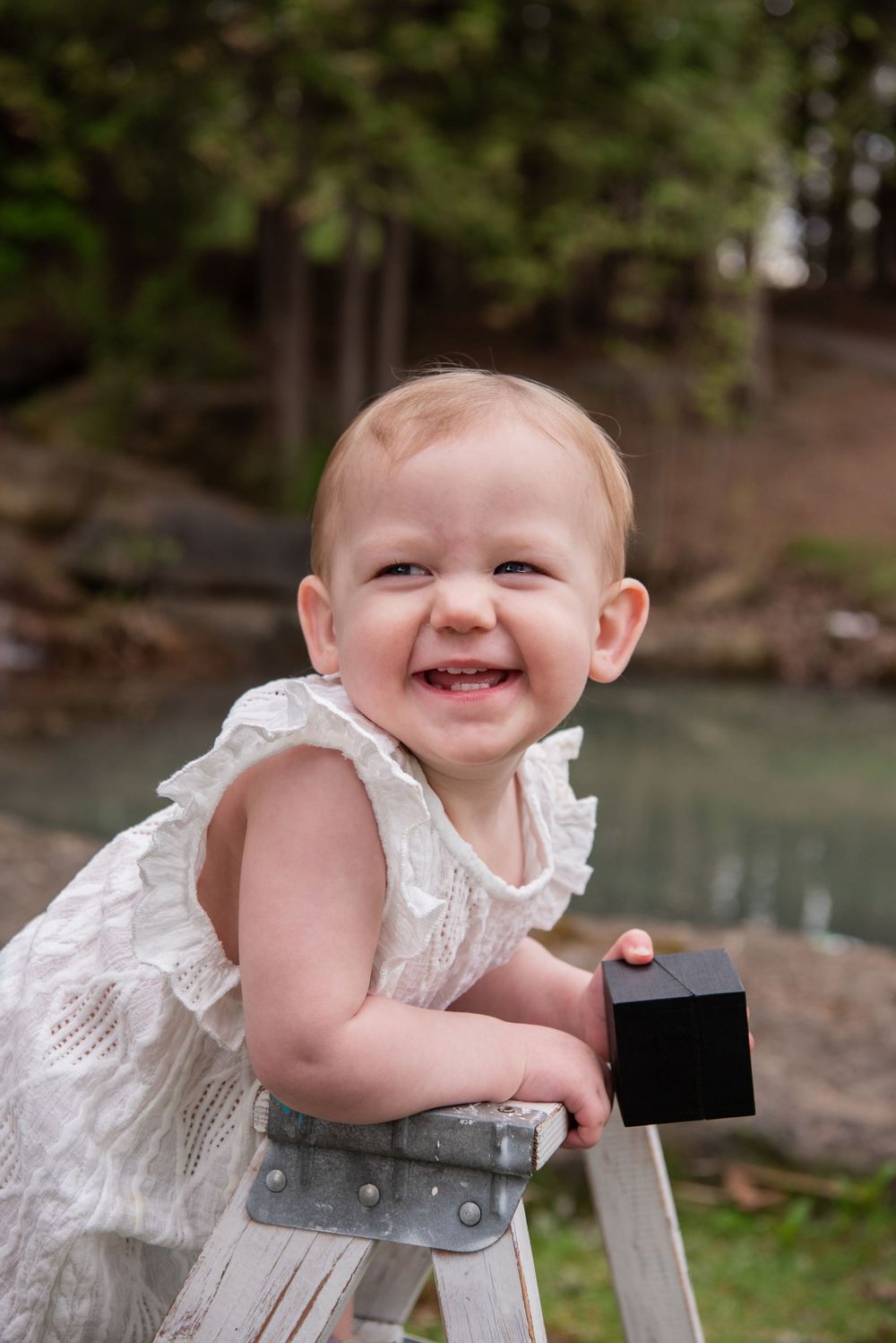 A toddler in a white lace dress smiles while standing on a wooden stool near a water feature.