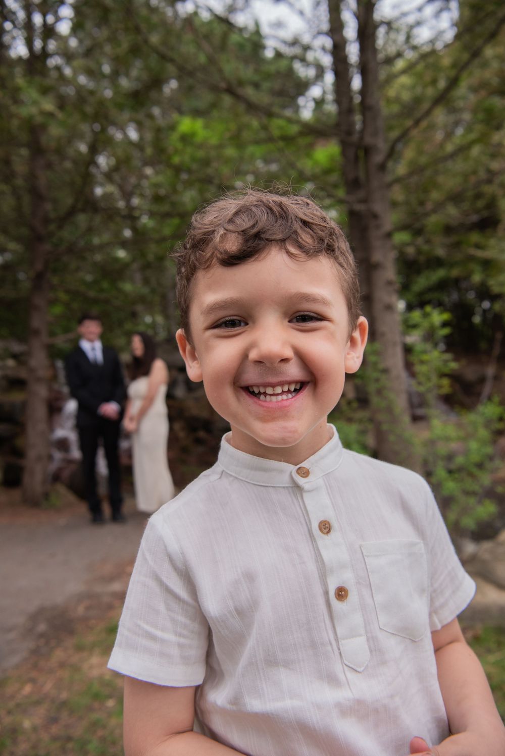 A child in a white linen shirt with buttons smiles joyfully in a forest setting with wedding guests visible in background.