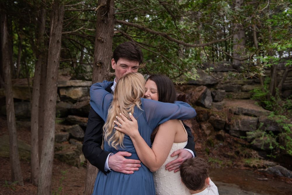 A group embrace in front of a stone wall in a wooded setting during what appears to be a special occasion.