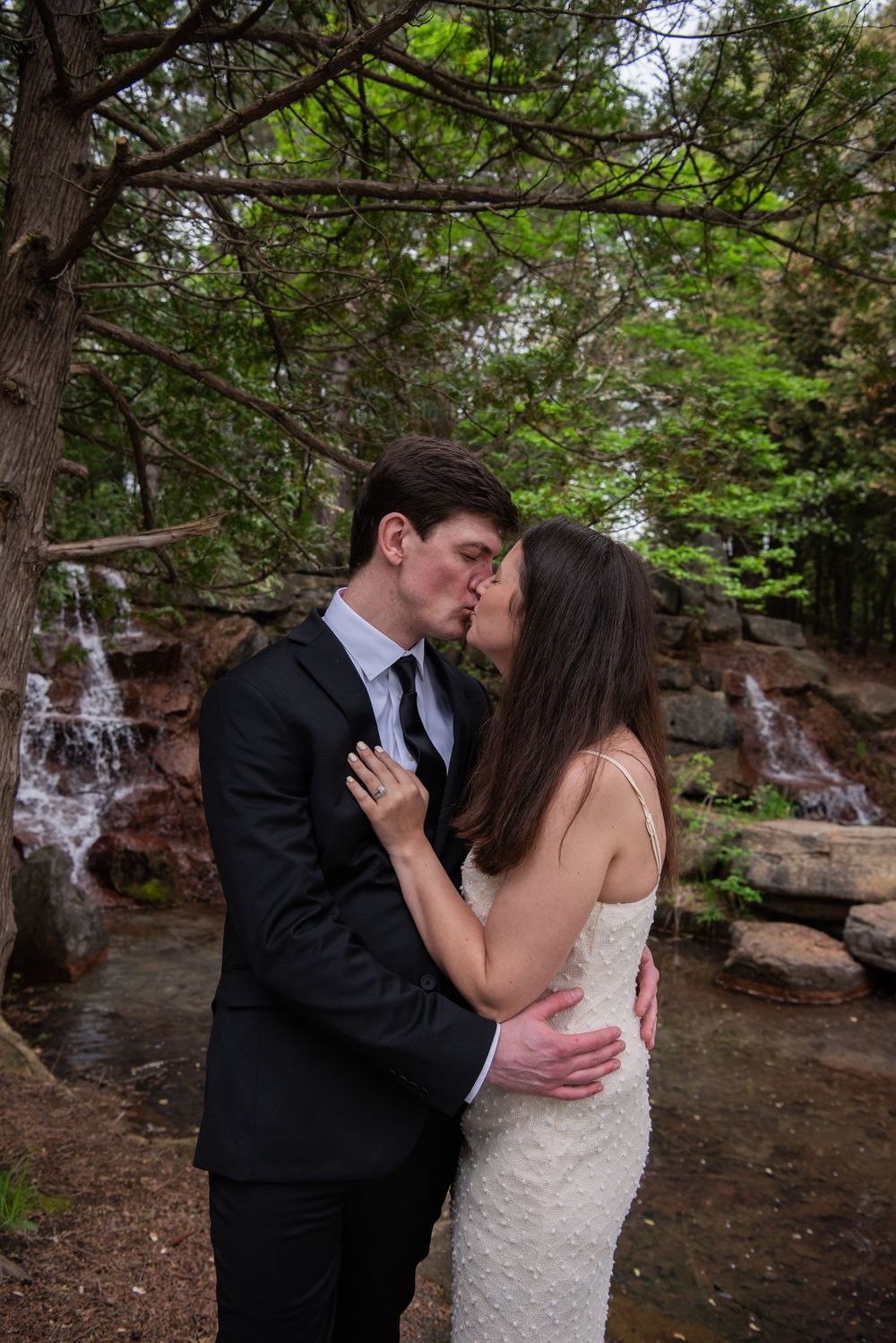 A romantic wedding couple shares a kiss by waterfalls in a lush forest setting.
